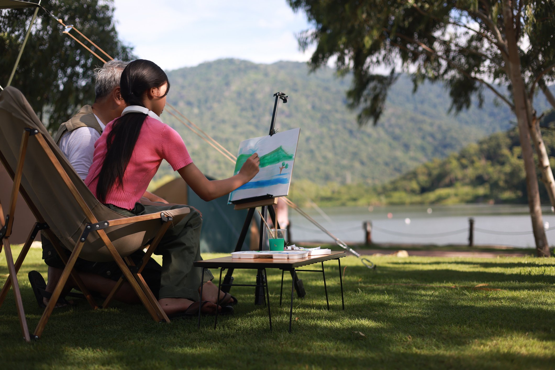 Seen from behind, a girl in a pink shirt sits in a folding chair, painting a landscape on an easel. Beside her sits an older man. They are outdoors by a lake with green mountains in the background. A small table holds art supplies next to a camping tent under large trees.