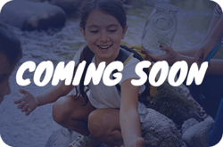A blue-tinted image of children exploring a stream, with the white text "COMING SOON" overlaid in the center. One child is smiling and reaching toward the water while another holds a glass jar.