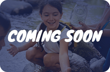A blue-tinted image of children exploring a stream, with the white text "COMING SOON" overlaid in the center. One child is smiling and reaching toward the water while another holds a glass jar.