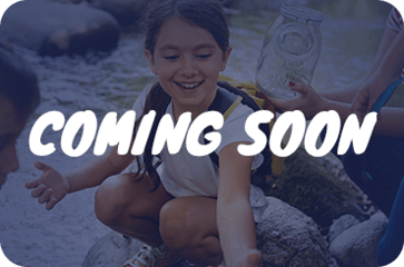 A blue-tinted image of children exploring a stream, with the white text "COMING SOON" overlaid in the center. One child is smiling and reaching toward the water while another holds a glass jar.