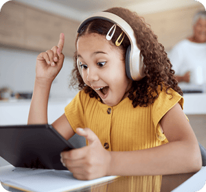 A young girl with curly hair and headphones looks at a tablet with an expression of pure joy and surprise. She points one finger upward in a "Eureka" gesture. She wears a yellow ribbed shirt and colorful hair clips. In the blurred background, an older woman smiles in a bright kitchen.