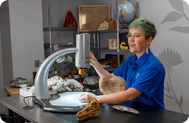 A woman with short green hair uses a silver digital microscope to examine fossils. An ammonite, a dinosaur skull, and a globe are on the desk. She wears a blue button-down shirt and looks toward the side with a slight smile. Shelves in the background hold various scientific specimens. A woman with short green hair uses a silver digital microscope to examine fossils. An ammonite, a dinosaur skull, and a globe are on the desk. She wears a blue button-down shirt and looks toward the side with a slight smile. Shelves in the background hold various scientific specimens.