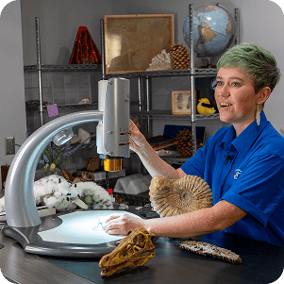 A woman with short green hair uses a silver digital microscope to examine fossils. An ammonite, a dinosaur skull, and a globe are on the desk. She wears a blue button-down shirt and looks toward the side with a slight smile. Shelves in the background hold various scientific specimens.
