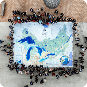 An aerial bird's-eye view shows a large group of people standing around the perimeter of a massive floor map of the Great Lakes and St. Lawrence River basin. The map is highly detailed with blue water and green land. The crowd is gathered on a light gray pavement next to a wooden deck.