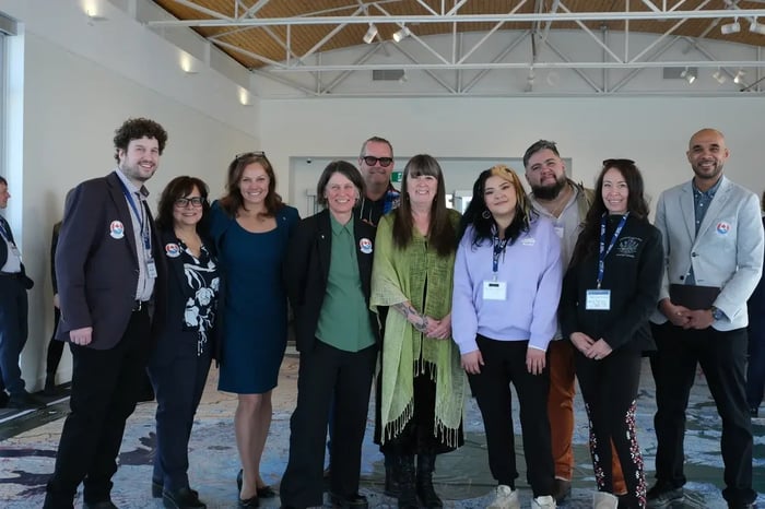 Ten representatives from AquaAction, the Canadian Museum of Nature, RCGS, and Roots of Resilience stand in a row on a large geographic floor map in a bright hall. They wear business-casual clothing with event pins and lanyards, smiling toward the camera.