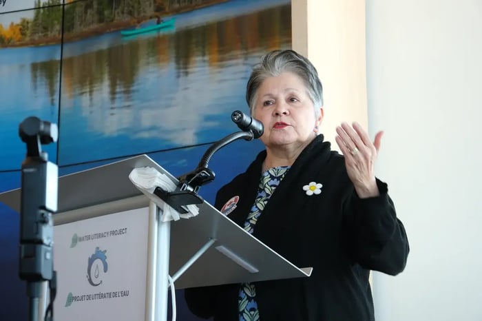 Claudette Commanda speaks into a microphone at a metal podium bearing "The Water Literacy Project" logo. Gesturing with her hand, she wears a black cardigan with a beaded daisy pin over a floral shirt. Behind her, a screen displays a canoe on a calm lake.