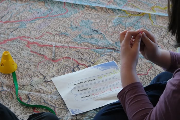 A student sits beside a large topographic floor map of Yukon. Next to them, a worksheet reads: "My water started here... at Aishihik", "It travelled to... Arctic Ocean", and "It helped... Tutchone du S...". A white crayon and a yellow plastic marker rest on the map.