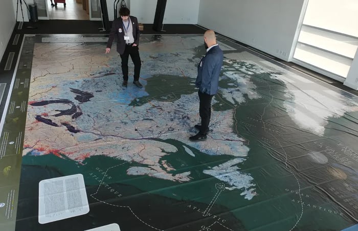 Two men in business attire stand on a giant vinyl floor map of Canada and North America inside a bright room. The man on the left points down toward a specific region while the other observes. The map displays detailed water bodies and bilingual labeling.