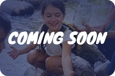 A blue-tinted image of children exploring a stream, with the white text "COMING SOON" overlaid in the center. One child is smiling and reaching toward the water while another holds a glass jar. A blue-tinted image of children exploring a stream, with the white text "COMING SOON" overlaid in the center. One child is smiling and reaching toward the water while another holds a glass jar.