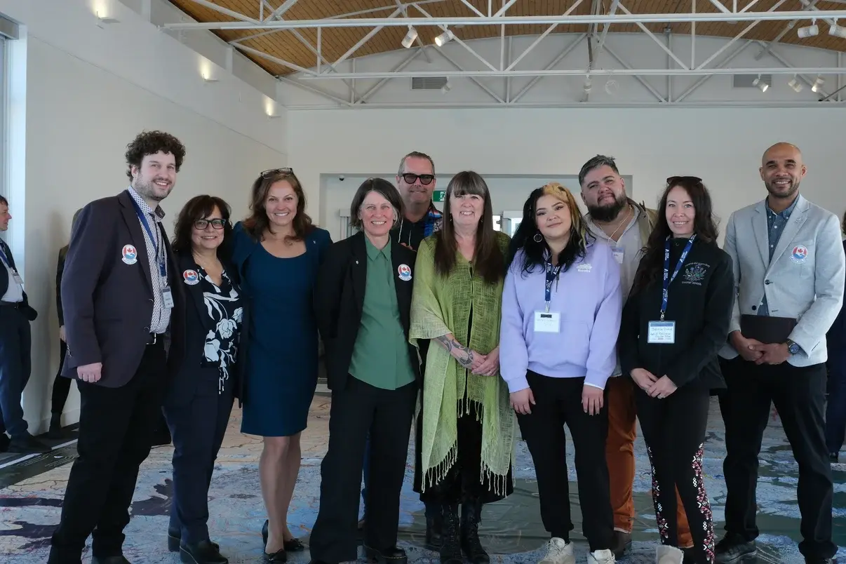 Ten representatives from AquaAction, the Canadian Museum of Nature, RCGS, and Roots of Resilience stand in a row on a large geographic floor map in a bright hall. They wear business-casual clothing with event pins and lanyards, smiling toward the camera.